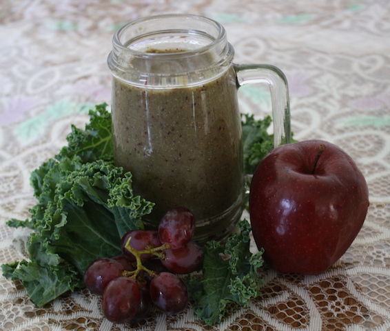 a green smoothie in a glass mug surrounded by torn Kale garnished with a whole red apple and purple grapes