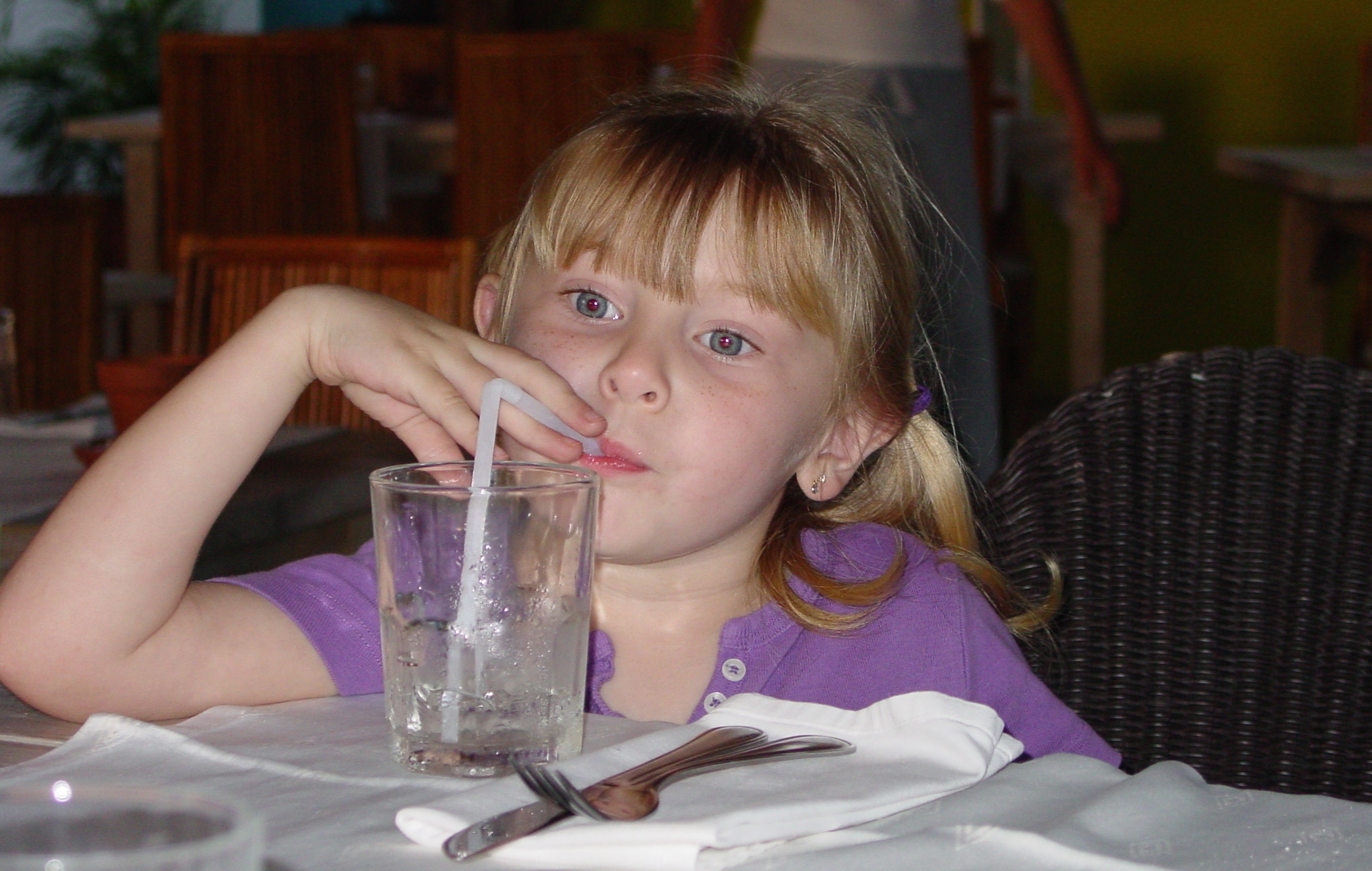 a cute little girl at the dinner table drinking a cool glass of water.
