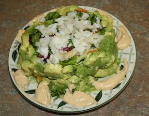 avocado salad with a ring of avocados surrounding a crispy filling of field greens topped with flaky cod with a little sauce on  the plate to use as you desire