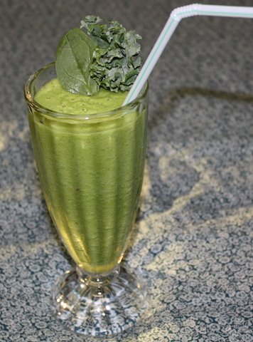 A tall fluted glass filled with a pretty green colored smoothie decorated with crinkly kale served with a straw.