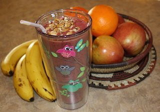 A bright rose colored smoothie topped with nuts sitting on the counter by some fruit makes you hungry